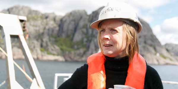 

En kvinne med oransje redningsvest og hvit hjelm står på dekk av et fartøy. Hun holder en kaffikopp i hånden og ser ut av bildet. Bak henne ligger Jøssingfjord.