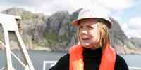 

En kvinne med oransje redningsvest og hvit hjelm står på dekk av et fartøy. Hun holder en kaffikopp i hånden og ser ut av bildet. Bak henne ligger Jøssingfjord.