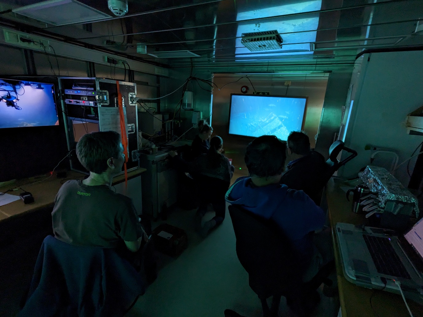 Photo of people with their backs to the camera in a dark room with all looking at a tv where there is live imagery of the shipwreck.