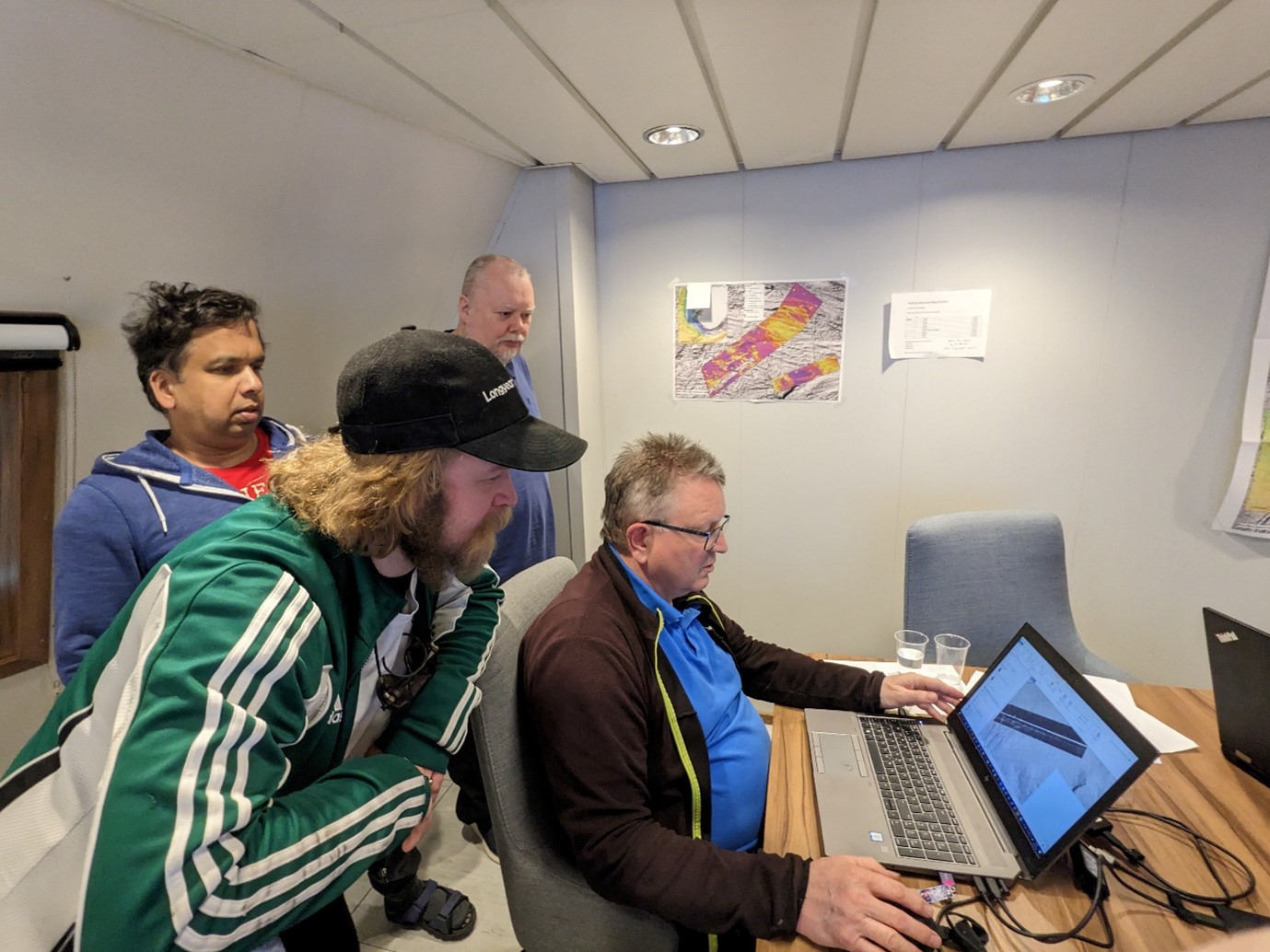 A photo of four people looking at a computer screen which is showing HiSAS bathymetry being mapped in a GIS