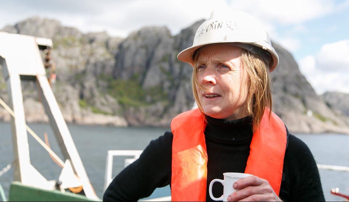 
En kvinne med oransje redningsvest og hvit hjelm står på dekk av et fartøy. Hun holder en kaffikopp i hånden og ser ut av bildet. Bak henne ligger Jøssingfjord.